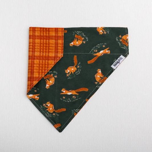 A triangular fabric pet bandana with a dark green background featuring orange foxes and an orange-brown plaid reverse side, laid flat on a white textured background. A white tag on the edge reads "Wendy & Co".