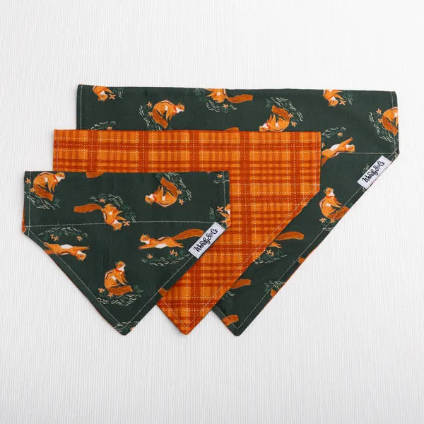 Three dog bandanas in a fan layout on a white textured background. The top bandana is dark green with an orange fox pattern, the middle bandana is orange and brown plaid, and the bottom bandana is the dark green with orange squirrel pattern showing its reverse side.