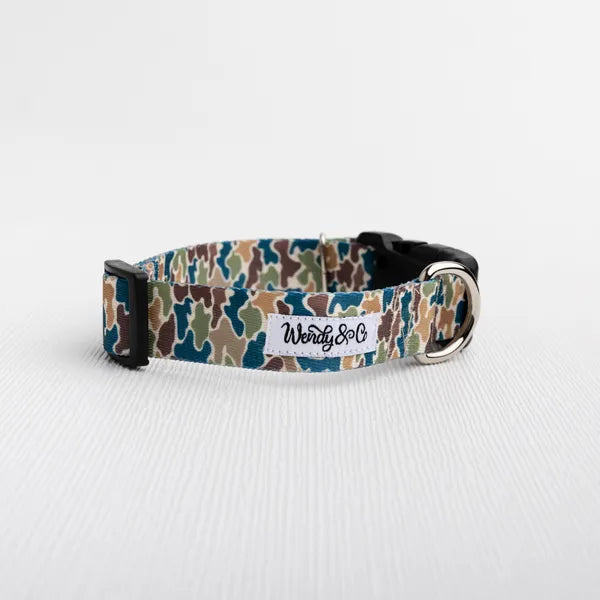 A camouflage dog collar with a blue, green, brown, and beige camo pattern, featuring a white brand label that reads 'Wendy&Co" and a black plastic buckle.