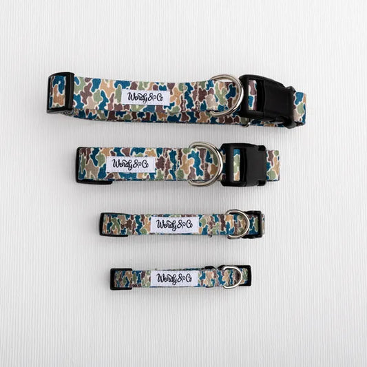 Four dog collars with a blue, green, and beige camouflage pattern, laid out vertically on a white textured background. Each collar has a white rectangular label with the brand name 'Wendy&Co' and a black plastic buckle.