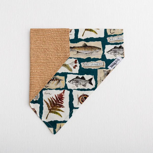 A square bandana-style pet collar or scarf laid flat on a white textured background. One side shows a warm tan fabric with small white dots, while the other side features a collage print with fish illustrations, fishing-related labels, trout and botanical details on a dark teal base.