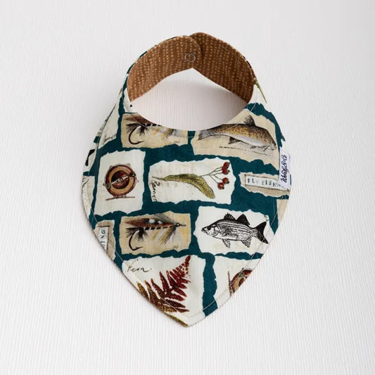A triangular bandana bib for dogs, featuring a vintage-style fish and nature print with trout, botanicals and fly fishing on a cream background with dark teal grid lines, displayed on a white textured background.