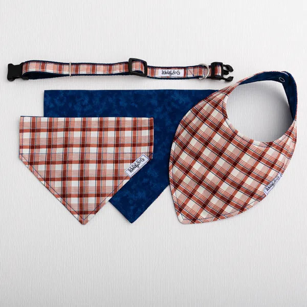 A pet accessory set laid flat on a white textured background, including a plaid bandana collar, a plaid collar, and a matching plaid bandana bib with a dark blue backing. 