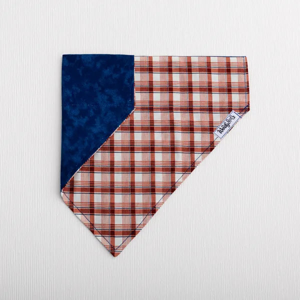 A small dog bandana laid flat on a white textured background. One corner is solid dark blue and main body feature a copper, cream and navy plaid pattern.