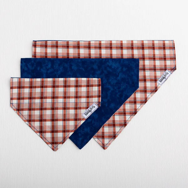 Three dog bandanas from the brand Wendy & co are arranged in a fan shape on a white textured background. The bandanas feature a navy blue reverse side and a copper, cream and navy plaid patterned front side.