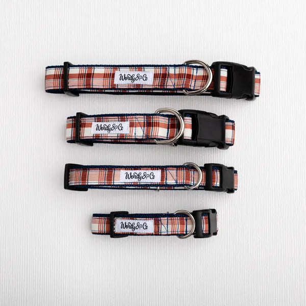 Three dog collars in different sizes with a red, white, and navy plaid pattern, black plastic buckles, and silver metal rings, laid out horizontally on a white textured background. 