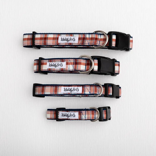Three dog collars in different sizes with a red, white, and navy plaid pattern, black plastic buckles, and silver metal rings, laid out horizontally on a white textured background. 