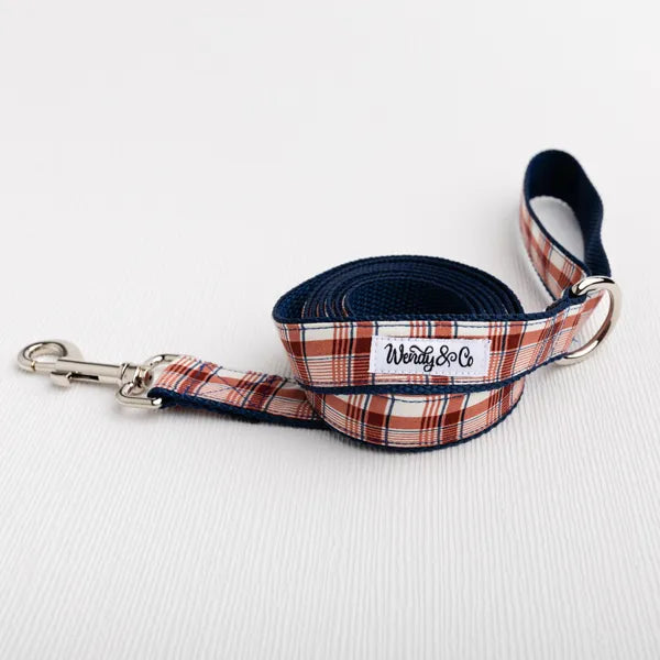 A plaid dog leash with copper, cream and blue plaid with silver metal hardware, coiled on a white textured surface. The brand label reads 'Wendy & Co".