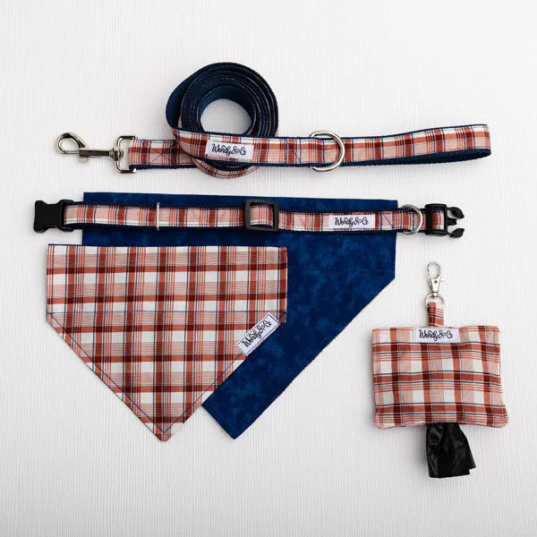 A pet accessory set laid out on a white textured background, including a matching leash, collar, bandana, and small poop bag. The bandanas and pouch feature a copper, cream, and navy plaid pattern, while the collar and leash have navy blue fabric with copper, cream and navy plaid detailing.