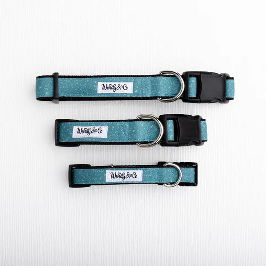 Three dog collars in a teal glitter fabric with black trim, metal D-rings, and black plastic buckles, laid out horizontally on a white textured background. 