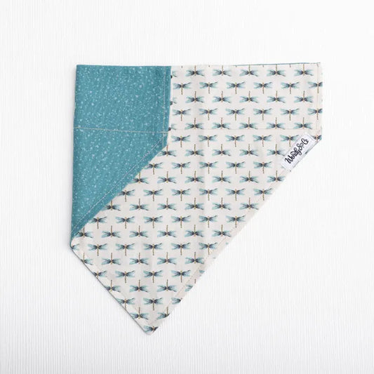 A triangular bandana-style pet collar or scarf laid flat on a white textured background. One half shows a teal fabric with tiny white speckles, and the other half shows an off-white background with a repeating teal dragonfly print.