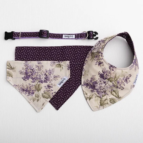 A pet accessory set laid flat on a white textured background, including a purple patterned collar, a dark purple bandana, a beige bandana with a purple floral print, and a matching beige bib with a purple floral print. 