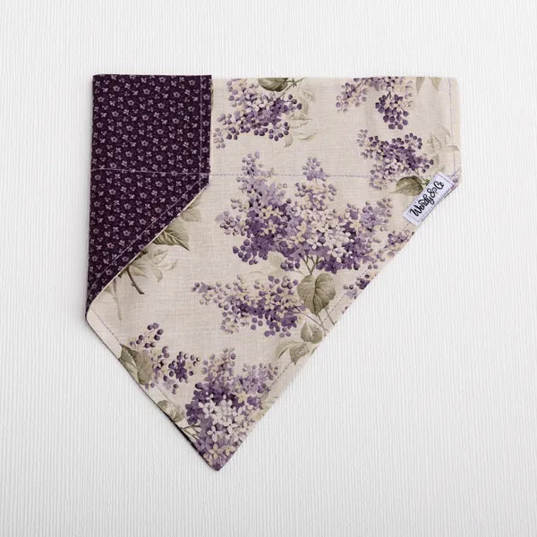 A triangular bandana-style pet collar or scarf laid flat on a white textured background. The fabric features a floral print in purple and beige with green leaves, and a solid dark purple patterned reverse side.