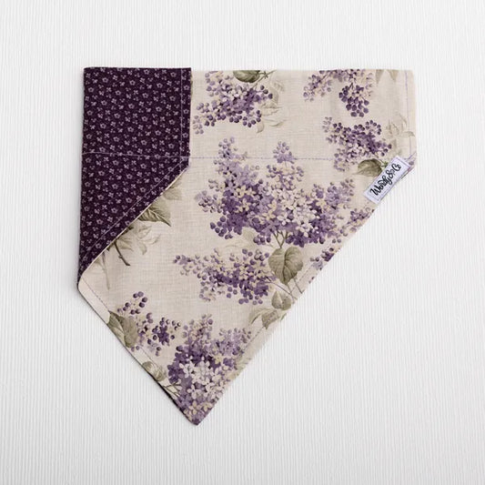 A triangular bandana-style pet collar or scarf laid flat on a white textured background. The fabric features a floral print in purple and beige with green leaves, and a solid dark purple patterned reverse side.