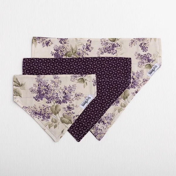 Three dog bandanas from the brand K&H Pet Supplies are arranged in a fan shape on a textured background. The top bandana features a light beige background with a purple floral print, the bottom-left bandana is solid dark purple, and the center bandana has a small purple floral pattern on a dark purple background.