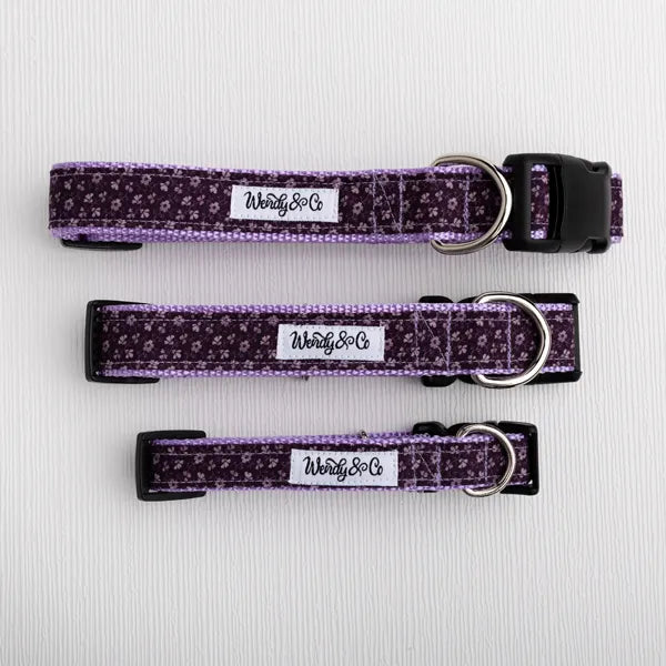 Three dog collars in a purple and lavender floral pattern with black plastic buckles and silver metal rings, laid flat on a white textured background. Each collar has a white fabric label with the brand name 'Wendyy & Co.