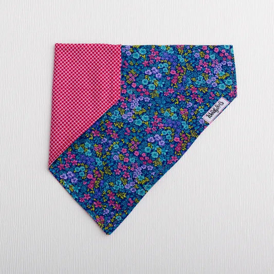 A triangular pet bandana with a split design: one half features a pink and white gingham pattern, while the other half displays a dense blue, purple, and pink floral print. A small white brand tag is visible on the floral side.
