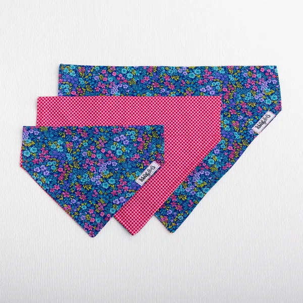 Three dog bandanas stacked on a white textured background. Two bandanas feature a blue, purple, and green floral pattern, and one is solid hot pink with a small red gingham check pattern.