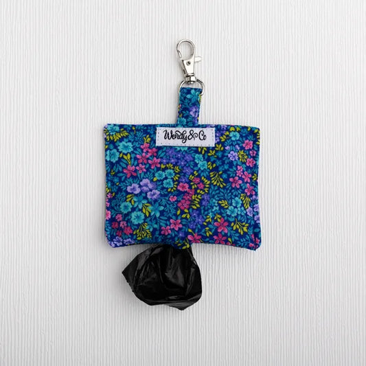A fabric dog poop bag holder with a floral print in blue, pink, and purple, featuring a silver clasp and a black plastic bag, displayed on a white textured background.
