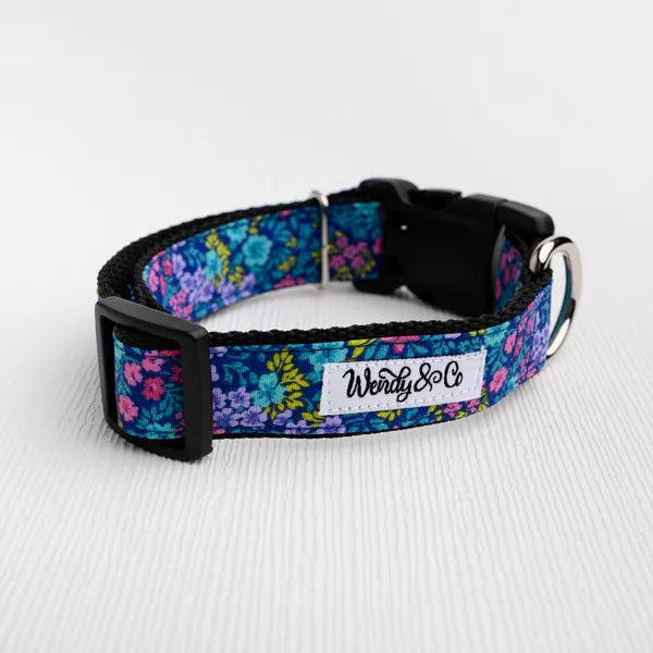 A dog collar with a vibrant blue, purple, and pink floral pattern, featuring a black plastic buckle and a white fabric label that reads 'Wendy & Co'.