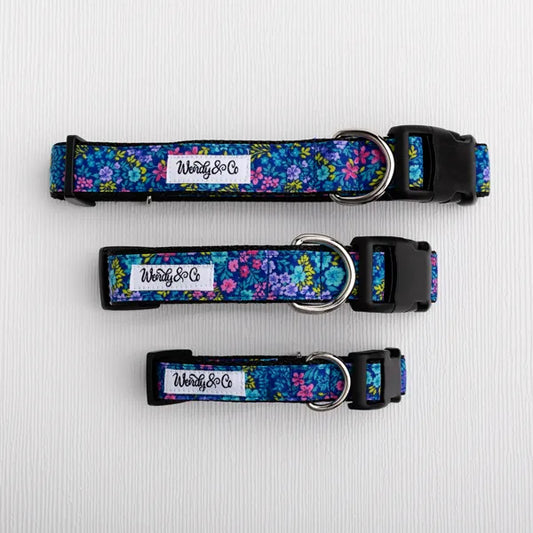 Three floral dog collars in different sizes arranged vertically on a white textured background. The collars feature a vibrant blue, purple, and pink floral pattern with black plastic buckles and silver metal rings.