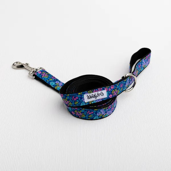 A coiled dog leash with a vibrant floral pattern in purple, blue, and pink, featuring a black underside, silver metal hardware, and a white brand label that reads 'Wendy & Co'.