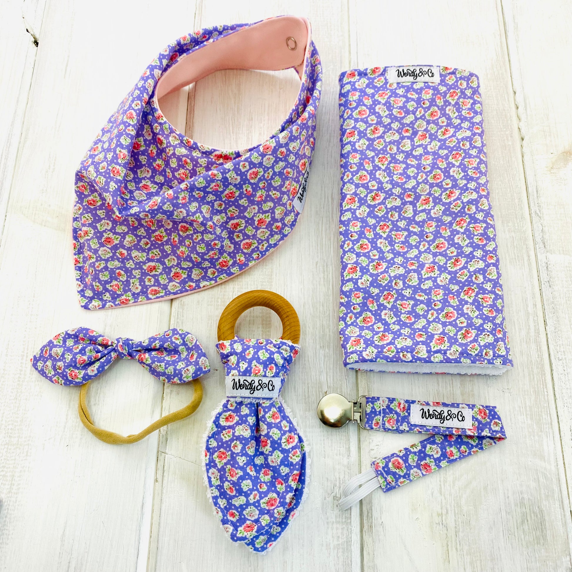 Baby shower gift set with bib, burp cloth, teether, paci clip and bow in a pastel lilac print.