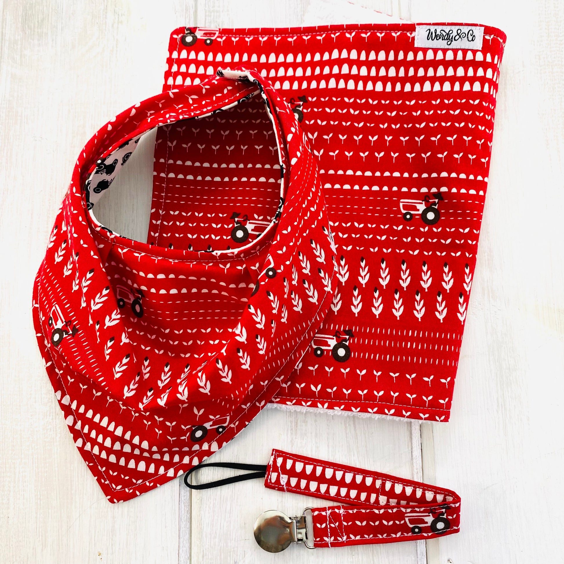 Bandana bib, pacifier clip and burp cloth in red, white and black fabric depicting a tractor plowing fields.
